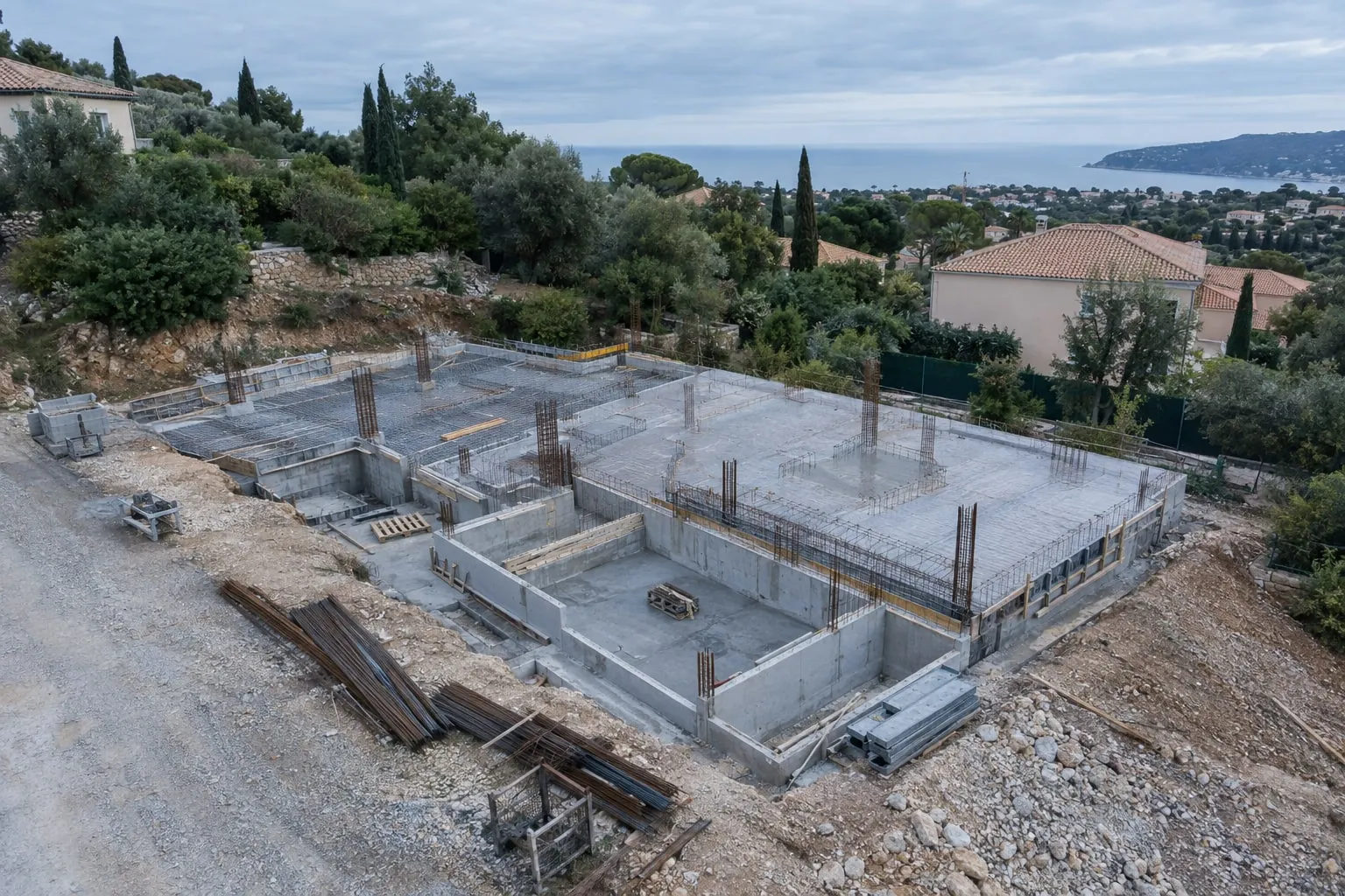 Chantier de gros œuvre vu du ciel, fondations et voiles béton d'une villa face à la mer