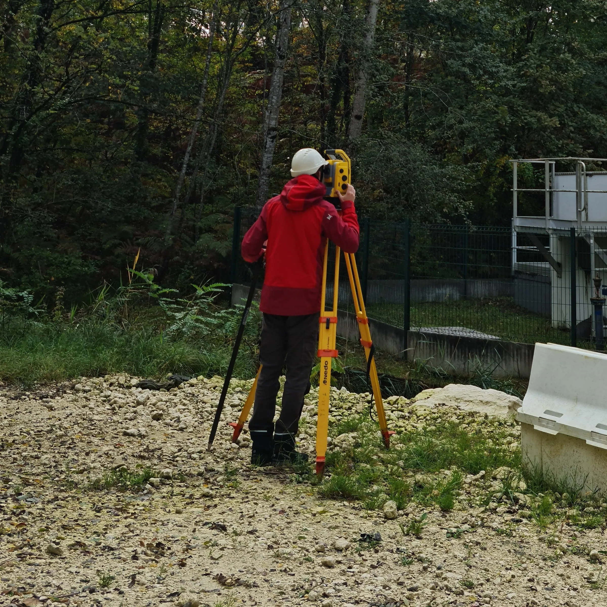 Géomètre topographe en veste rouge derrière une station totale, devant une station de pompage en lisière de forêt